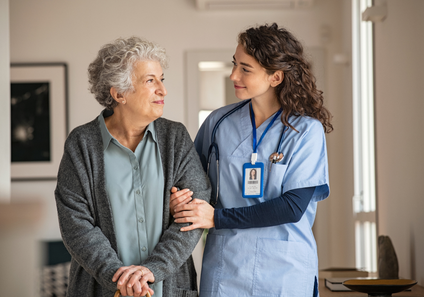 From left to right, a senior citizen is walking through a sunlit hallway with the support and assistance of a healthcare worker. Support for one another makes us all stronger. | RadarFirst