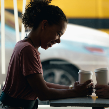 A young person converses with a friend at a coffee shop. The image foreground is dark, in shadows while the background illuminates a busy urban street. | RadarFirst